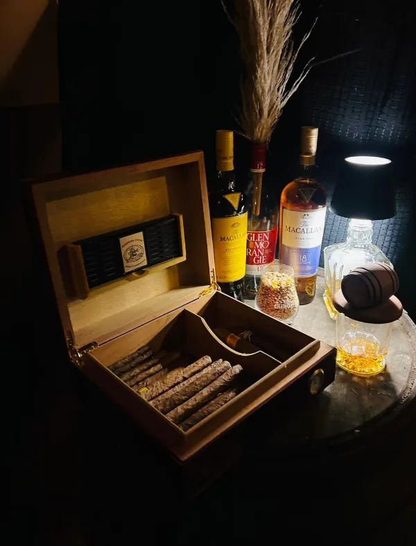 Caja de puros abierta junto a botellas de whisky y una lámpara de mesa