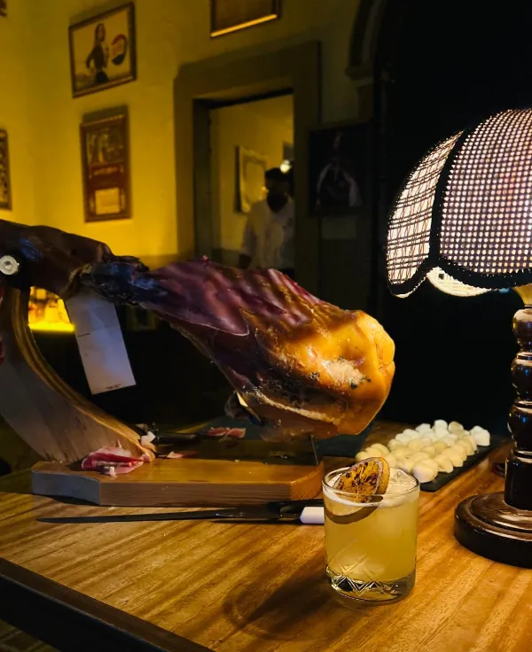 Pierna de jamón serrano en su base, junto a un cóctel con rodaja de cítrico tostada y una lámpara de mesa encendida