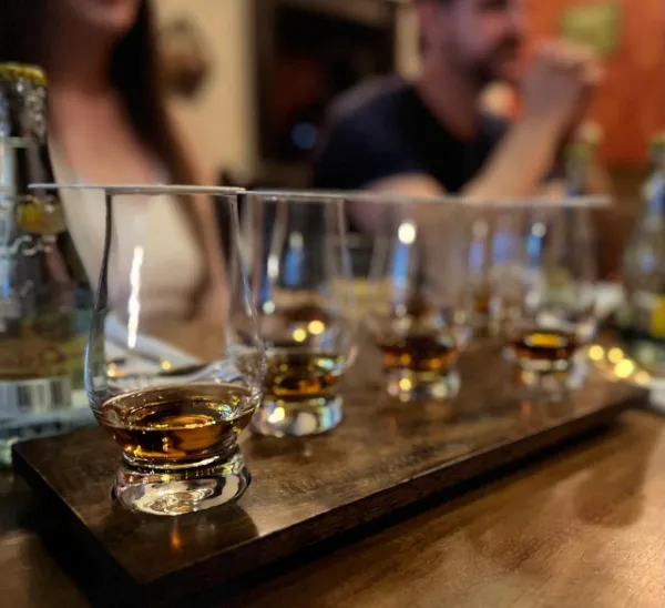 Tabla con una fila de copas de tequila para catar, tapadas con posavasos; al fondo, invitados conversando