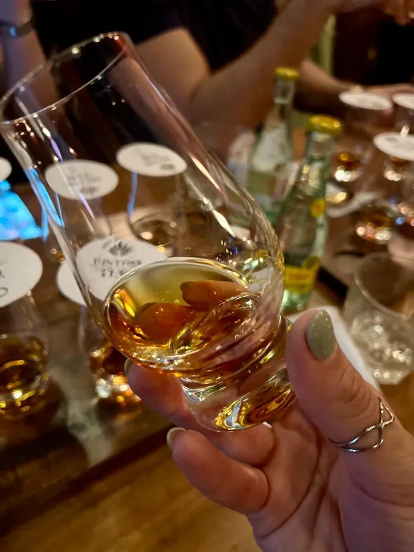 Una mano sostiene a contraluz una copa de tequila ambarino; atrás, las demás copas de la cata